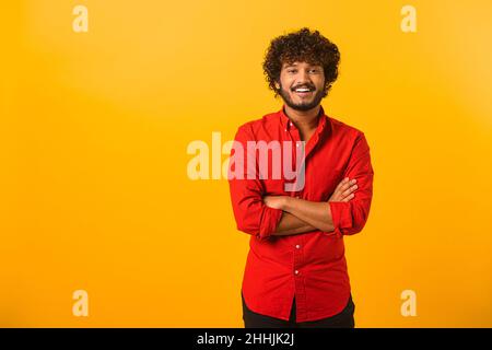 Ritratto di felice soddisfatto bel giovane uomo in camicia in piedi con le braccia incrociate e guardando la macchina fotografica con il sorriso toothy. Studio interno girato su sfondo arancione Foto Stock