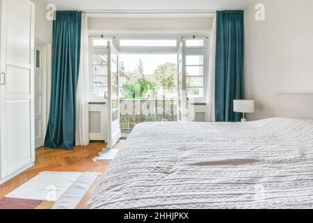 Comodo letto con coperta bianca posto vicino alla lampada e armadio spaziosa camera da letto con porta chiusa e grandi finestre a casa Foto Stock