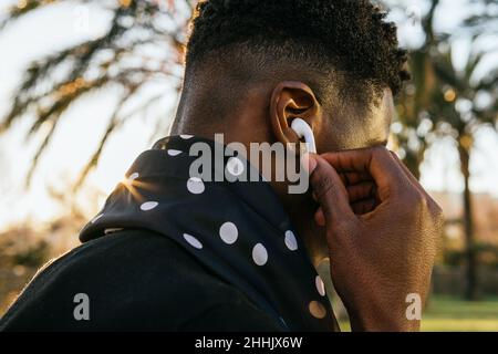 Vista laterale del tipo nero anonimo che regola i veri auricolari wireless durante l'ascolto della musica nel parco Foto Stock