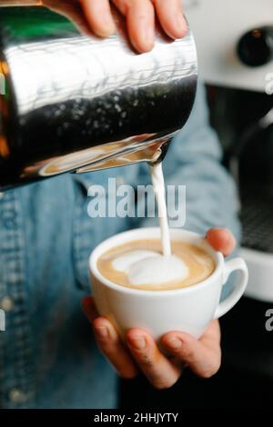 Ritagliare un barista irriconoscibile versando il latte dalla caraffa in una tazza di caffè appena fatto mentre si prepara il latte durante il lavoro in un caffè Foto Stock