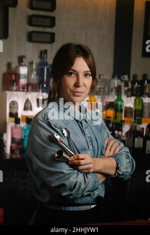 Attraente barista femminile con jigger e tong in mano guardando la macchina fotografica mentre si trova nel bar con varie bevande alcoliche Foto Stock