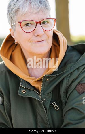 Crop grigio capelli senior donna in occhiali indossare outerwear caldo delicatamente sorridente alla fotocamera Foto Stock