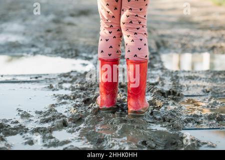 Taglia il bambino irriconoscibile in pantaloni rosa e stivali di gomma rossa in piedi su un sentiero sporco con le pozzanghere sulla strada della città Foto Stock