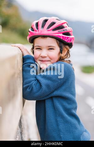 Ragazza felice carina in abbigliamento casual e casco protettivo rosa guardando la macchina fotografica mentre si trova vicino alla pietra recinzione sulla strada contro sfondo sfocato Foto Stock