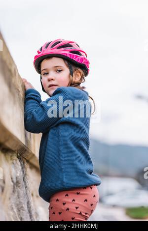 Ragazza carina in abbigliamento casual e casco protettivo rosa guardando la macchina fotografica mentre si trova vicino alla pietra recinzione sulla strada contro sfondo sfocato Foto Stock