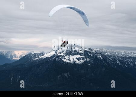 Vista aerea anonima di chi vola su parapendio bianco su un piccolo insediamento situato vicino a alte montagne rocciose coperte di neve nella città di Salisburgo Foto Stock