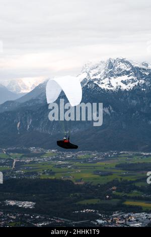 Vista aerea anonima di chi vola su parapendio bianco su un piccolo insediamento situato vicino a alte montagne rocciose coperte di neve nella città di Salisburgo Foto Stock