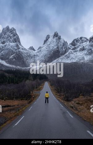 Lontano anonimo turista in piedi su strada asfaltata vuota contro le cime di montagna coperte di neve durante il viaggio in Norvegia il giorno d'inverno Foto Stock