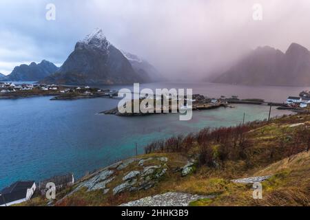 Da sopra di magnifica vista di montagne innevate nella nebbia e villaggi con case situate sul terreno erboso della baia nelle isole Lofoten in Norvegia Foto Stock