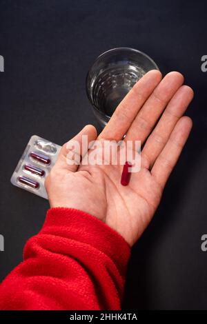 Da sopra di raccolto anonimo persona che mostra capsula rossa in mano sullo sfondo della tavola con un bicchiere d'acqua Foto Stock