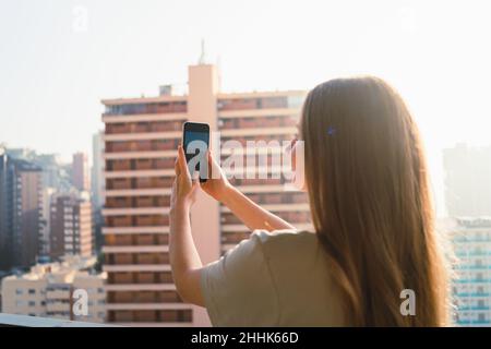 Positiva giovane viaggiatore femminile con capelli lunghi in abiti casual scattando foto su telefono cellulare in piedi sulla terrazza in moderno edificio a più piani in cit Foto Stock