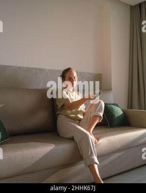 Calma giovane signora barrefooted in abiti casual che cambiano canali con telecomando mentre riposano da solo su un comodo divano in appartamento moderno in Foto Stock