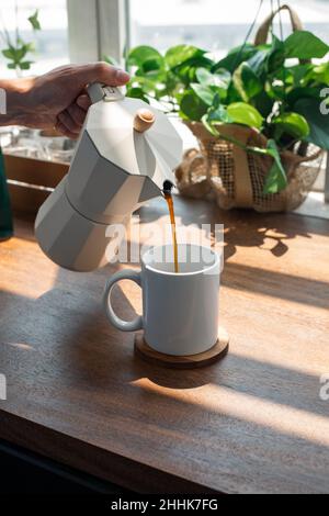 Ritagliare una persona irriconoscibile versando caffè caldo da una pentola di moka in metallo in una tazza bianca su un tavolo di legno in una cucina leggera a casa Foto Stock