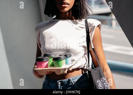 Crop African American femmina in abiti casual che trasportano cartoni con tazze di caffè da asporto in giornata di sole in città Foto Stock