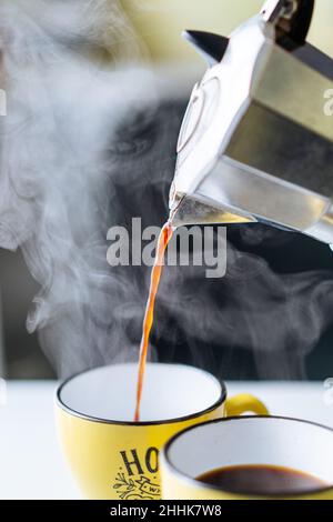 Caffè caldo appena fatto che si versa da una pentola di moka in metallo in una tazza gialla sul tavolo in una cucina chiara su sfondo sfocato Foto Stock