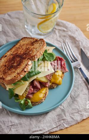 Da sopra di appetitoso sandwich con uova di carne e formaggio decorare con erbe in pane tostato servito su piastra di ceramica su tavola di legno con bicchiere di W. Foto Stock