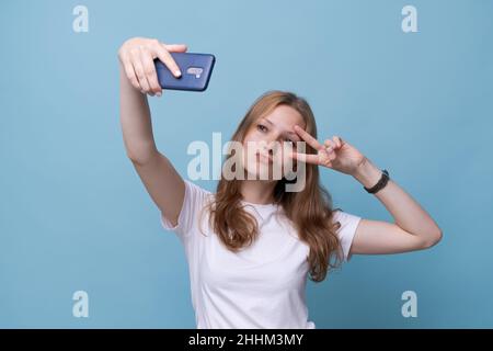 Ritratto felice amichevole giovane caucasica ragazza in bianco t-shirt su sfondo blu prendendo selfie e la mano ondeggiante, videochiamata, chat online. Il concetto di comunicazione tramite Internet Foto Stock