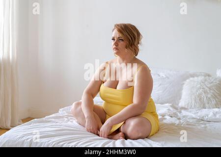 Modello Plus-size in abito giallo slinky siede sul letto accanto alla sua camera da letto, contemplando il suo aspetto. La giovane donna sorridente decide cosa indossare. XXL moda Foto Stock