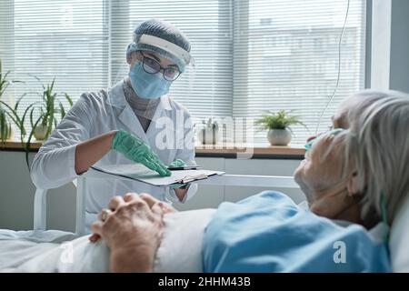Giovane dottore in abbigliamento protettivo che spiega i metodi di trattamento per la donna anziana che si trova a letto al reparto Foto Stock