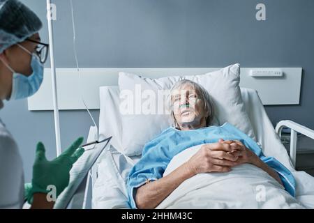 Donna anziana che parla con il suo medico in abbigliamento protettivo, che discute di trattamento mentre si trova in ospedale reparto Foto Stock