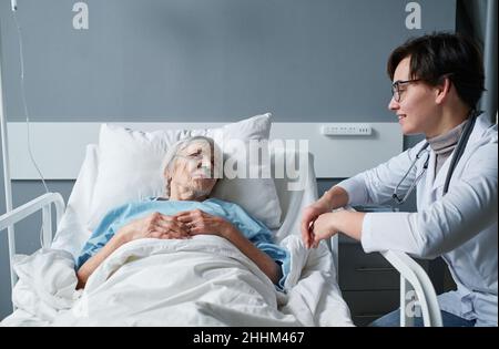 Donna anziana malata che riposa sul letto e parla con l'infermiera che si prende cura di lei in ospedale Foto Stock