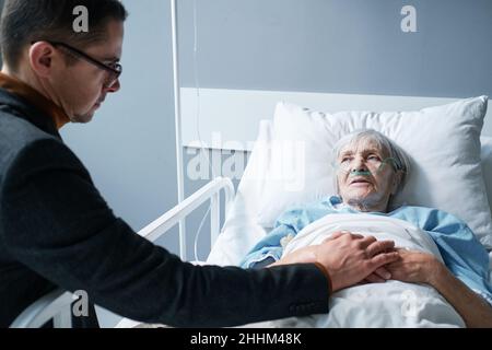 Madre anziana malata che si trova a letto nel reparto ospedaliero e parla con il figlio mentre tiene la mano Foto Stock