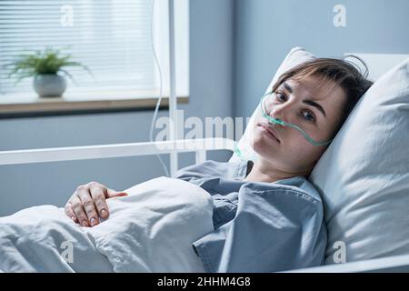 Ritratto di giovane donna sdraiata a letto con tubo di ossigeno guardando la macchina fotografica mentre si tratta in ospedale Foto Stock