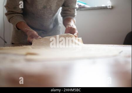 Vista ad angolo basso di una donna che prepara pasta di pasticceria vegana fatta in casa, tirandola e stirandola su un tavolo da pranzo domestico. Foto Stock