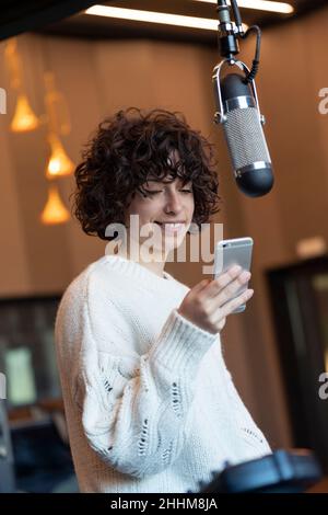 Caucasica giovane capelli ricci cantante donna sorridente con il suo telefono in uno studio di musica Foto Stock