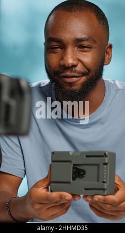 Influencer che esamina la batteria di alimentazione con montaggio a V sulla telecamera per consigli sul canale. Vlogger filma video per rivedere il caricatore portatile ricaricabile v lock, utilizzando treppiede e attrezzatura. Foto Stock