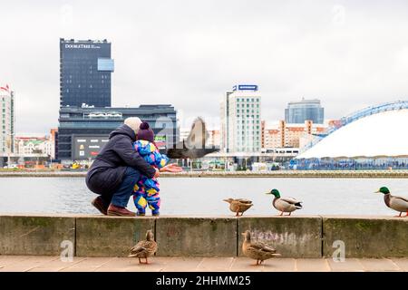 Madre e bambino nutrono uccelli sul lungofiume di Svisloch a Minsk. Sullo sfondo si trova l'hotel DoubleTree by Hilton e un centro commerciale Foto Stock