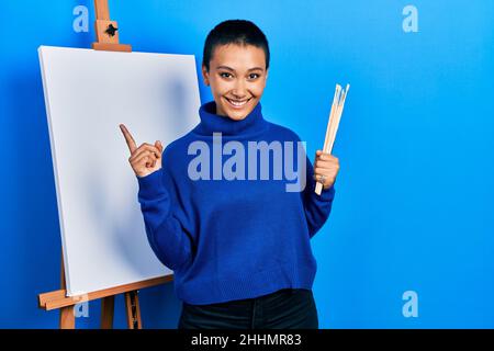 Bella donna ispanica con i capelli corti che tengono le spazzole vicino al cavalletto sorridendo felice di puntare con la mano e il dito al lato Foto Stock