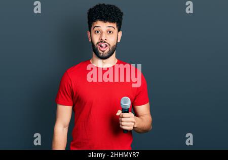 Giovane uomo arabo con la barba cantare canzone utilizzando microfono spaventato e stupito con bocca aperta per sorpresa, incredulità volto Foto Stock