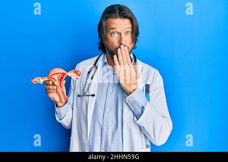 Uomo ginecologo di mezza età bello che tiene il modello anatomico di organo genitale femminile che copre la bocca con la mano, scioccato e impaurito per errore Foto Stock