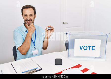 Uomo di mezza età con barba seduta da scrutinio tenendo io voto badge guardando fiducioso alla macchina fotografica sorridendo con le braccia incrociate e la mano sollevata sul mento Foto Stock