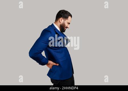 Ritratto della vista laterale dell'uomo barbuto in piedi con gli occhi chiusi, che soffre di dolore alla schiena più basso, infiammazione renale, vestito di stile ufficiale. Studio interno girato isolato su sfondo grigio. Foto Stock