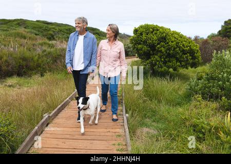 Felice coppia caucasica matura guardare via mentre si cammina con il cane sul lungomare. Insieme, week-end e natura concetto. Foto Stock