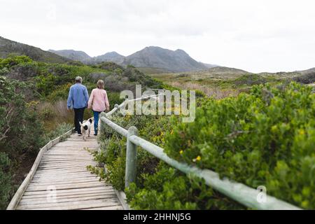 Vista posteriore della coppia caucasica che cammina con il cane sul lungomare durante il fine settimana. Concetto di convivenza, fine settimana e natura. Foto Stock