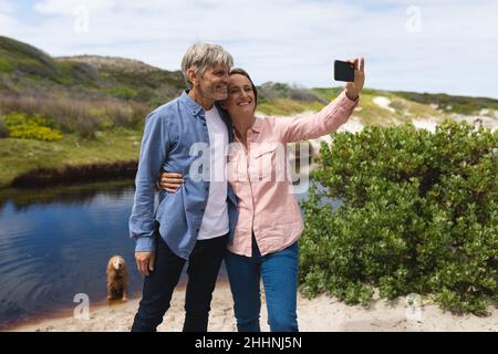 Felice coppia caucasica matura prendere selfie attraverso il telefono cellulare a Lakeshore durante il fine settimana. Insieme, week-end, wireless tecnologia e natura co Foto Stock