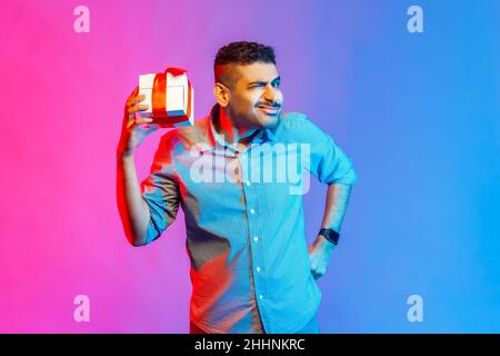 Ritratto di uomo divertente in camicia che tiene in mano la scatola presente, essendo interessato che cosa all'interno, volto accigliato, ha espressione insidiosa. Studio interno girato isolato su colorato sfondo di luce al neon. Foto Stock
