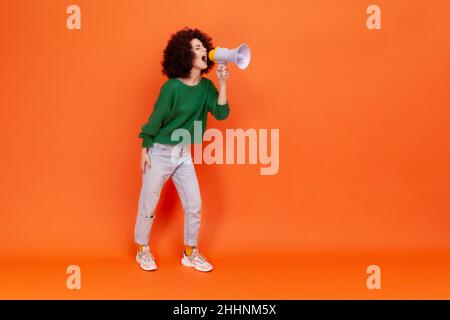 Ritratto a tutta lunghezza di donna con acconciatura Afro indossando un maglione verde casual in piedi urlando forte nel megafono, protestando. Studio interno girato isolato su sfondo arancione. Foto Stock