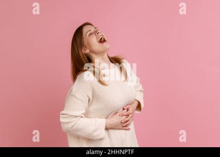 Ritratto di felice donna bionda che tiene il ventre, ridendo fuori forte, brulicante con risate isteriche, scherzando amico, indossando il maglione bianco. Studio interno girato isolato su sfondo rosa. Foto Stock