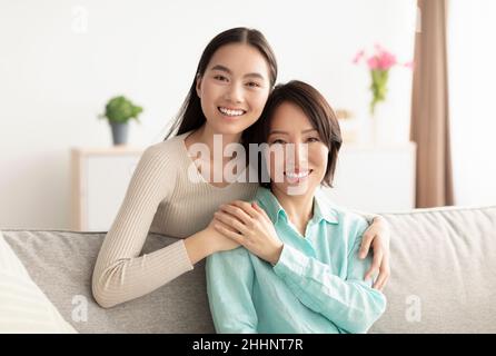 Felice donna asiatica matura e sua figlia adulta abbracciando e sorridendo a macchina fotografica, seduto sul divano a casa Foto Stock