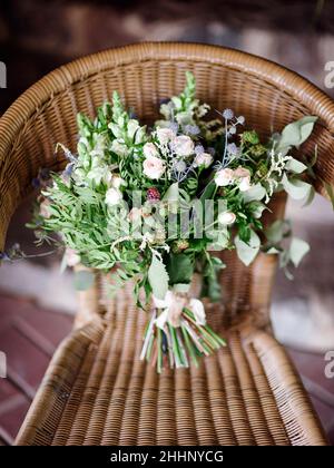 Bouquet di nozze su una sedia in vimini. Mazzo estivo con fiori di cardo blu, rose di jana, eucalipto e foglie d'oliva, bacche nere, snapdragon bianco fl Foto Stock