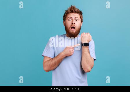 Ritratto di uomo sconvolto e spaventato in piedi con bocca aperta e meravigliato espressione, puntando con dito smartwatch ar. Studio interno girato isolato su sfondo blu. Foto Stock