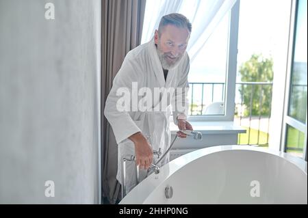 Uomo che si appoggia sulla vasca da bagno e accende il rubinetto Foto Stock