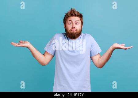 Così chi si preoccupa. Ritratto di uomo confuso bearded diffonde palme con esitazione non ha indizio o idea si alza perplesso, guardando impotente alla fotocamera. Studio interno girato isolato su sfondo blu. Foto Stock