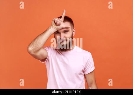 Ritratto di uomo bearded che tiene la mano vicino alla fronte mostrando il gesto perdente con le dita, avendo problemi finanziari e di affari, indossando la T-shirt rosa. Studio interno girato isolato su sfondo arancione. Foto Stock