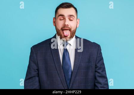 Ritratto di bambino spensierato uomo bruna indossare tuta stile ufficiale mostrando la lingua e gli occhi di chiusura con naughty disobbediente grimace, facendo il viso. Studio interno girato isolato su sfondo blu Foto Stock
