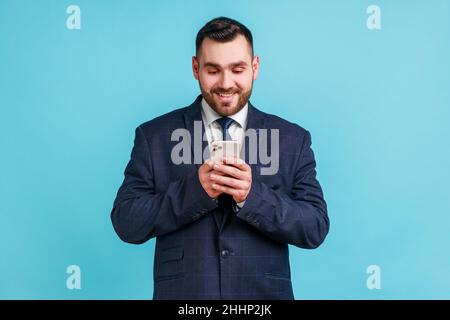 Ritratto di uomo d'affari allegro indossare vestito di stile ufficiale utilizzando il cellulare e sorridente, lettura di buone notizie messaggio, godendo applicazione mobile. Studio interno girato isolato su sfondo blu. Foto Stock